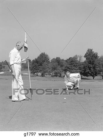 Stock Photo - 1930S 1940S 2 Elderly Men On Golf Green One Holds Flag And The Other Kneels Lining Up His Putt . Fotosearch
