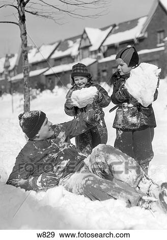 Stock Photo - 1930S 1940S Man Father Two Children Playing In Winter Snow Father Lying Down In Snow Kids Hold Huge Snow Balls Fun. Fotosearch