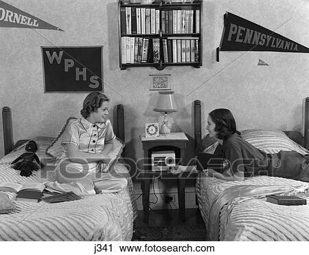Stock Image - 1930S 1940S Two Teen Girls Lying On Dormitory Beds Room Mates Listening To Radio College School Pennants On Wall Dorm. Fotosearch