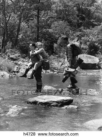 Stock Photo - 1930S 3 People Crossing Fording Stream One Man Carries Woman Piggy Back Other Carries Angling Fishing Gear Stepping Stones Water. Fotosearch