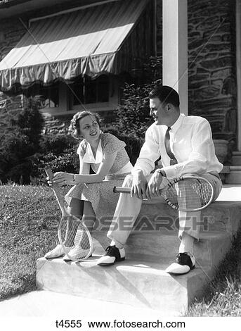 1930S Couple Holding Tennis Rackets Woman In Dress Man In Tennis Whites And Saddle Shoes Sitting On Steps Of House Smiling Talk View Large Photo Image Stock Photography - 1930S Couple Holding Tennis Rackets Woman In Dress Man In Tennis Whites And Saddle Shoes Sitting On Steps Of House Smiling Talk. Fotosearch