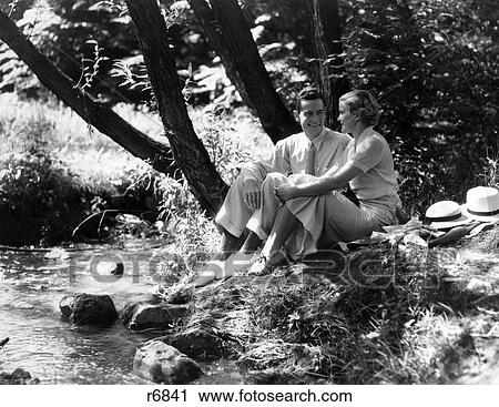 Stock Image - 1930S Couple Man Woman Sitting Beside Stream Side By Side Talking Flirting Date Dating. Fotosearch