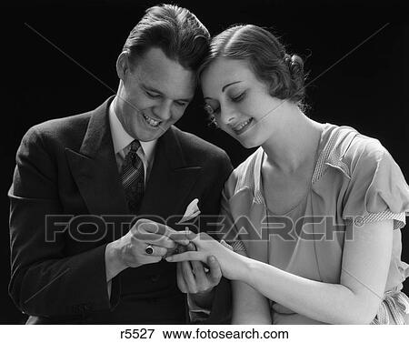 1930S Couple Sitting Next To Each Other With Heads Pressed Together Man Putting Engagement Ring On Woman'S Finger View Large Photo Image Stock Photo - 1930S Couple Sitting Next To Each Other With Heads Pressed Together Man Putting Engagement Ring On Woman'S Finger . Fotosearch