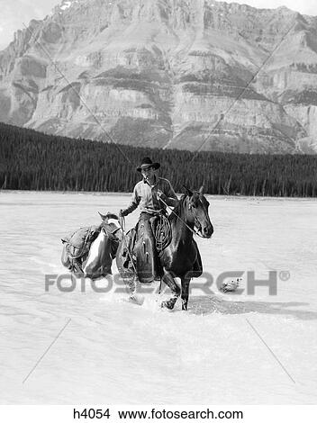 1930S Cowboy With Batwing Chaps On A Bay Horse Crossing A River Leading A Paint Pack Horse WithMountains In Background View Large Photo Image Picture - 1930S Cowboy With Batwing Chaps On A Bay Horse Crossing A River Leading A Paint Pack Horse WithMountains In Background . Fotosearch