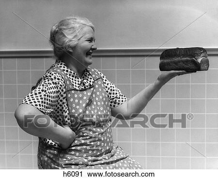 Stock Image - 1930S Grandmother In Apron In Kitchen Admiring Loaf Of Freshly Baked Bread . Fotosearch