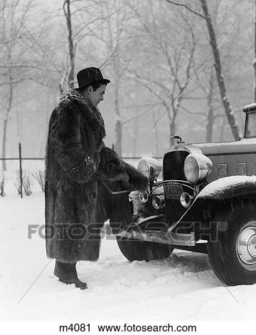 Stock Image - 1930S Man In Fur Coat & Hat Cranking Chevrolet In Snow . Fotosearch
