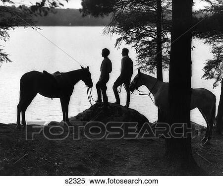 Stock Photography - 1930S Silhouette Of Couple Standing On Mound At Waterfront Holding Reins Of Horses To Either Side . Fotosearch
