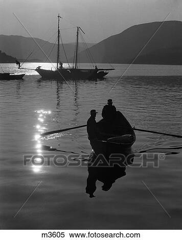 Stock Photography - 1930S Silhouette Of Pair Of Men In Rowboat With Sailboat In Background & Ray Of Sun Reflected On Water . Fotosearch