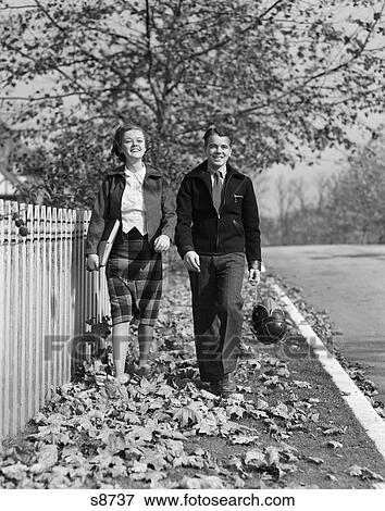 Stock Photo - 1930S Teen Couple Walking On Sidewalk In Fall Girl Carrying Schoolbooks Boy Carrying Football And Leather Helmet Smiling Outdoor. Fotosearch