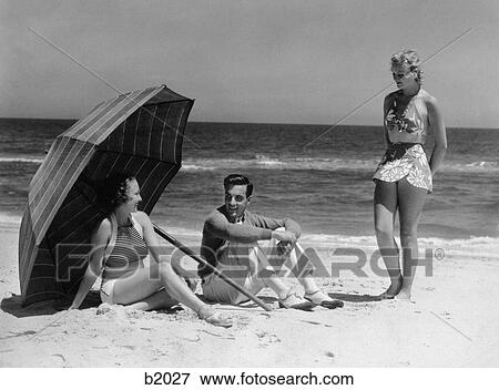 1930S Two Women 1 Man Sitting Under Beach Umbrella For Shade Wearing Fashionable Swim Wear View Large Photo Image Stock Photo - 1930S Two Women 1 Man Sitting Under Beach Umbrella For Shade Wearing Fashionable Swim Wear . Fotosearch