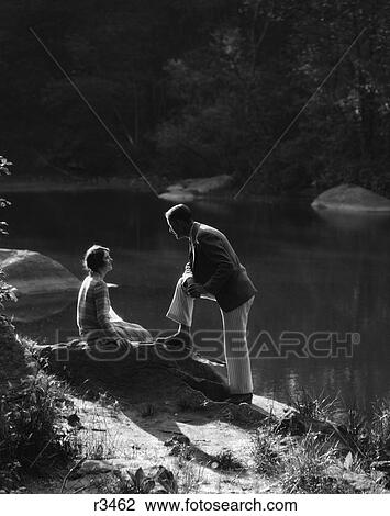 1930S Woman Sitting On Rock Lakeside Talking To Man Standing With Foot On Rock Leaning On Knee To Listen To Her View Large Photo Image Stock Image - 1930S Woman Sitting On Rock Lakeside Talking To Man Standing With Foot On Rock Leaning On Knee To Listen To Her . Fotosearch