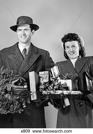 Stock Photo - 1940 1940S Couple Standing In Winter Coats Holding Wrapped Gifts And A Wreath Studio . Fotosearch