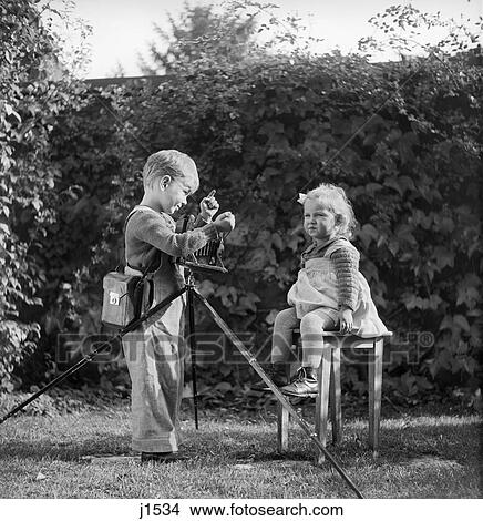 1940S 1950S Boy With Camera On Tripod Taking Photograph Of Baby Girl Toddler Sitting On Stool Director Pose View Large Photo Image Picture - 1940S 1950S Boy With Camera On Tripod Taking Photograph Of Baby Girl Toddler Sitting On Stool Director Pose . Fotosearch