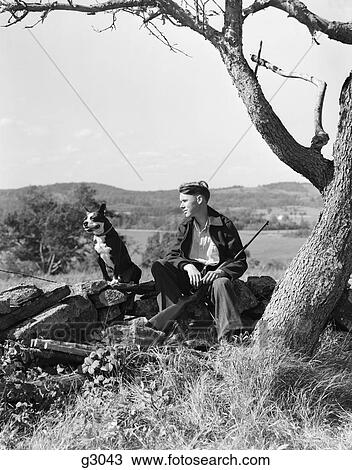 Stock Image - 1940S Boy Sitting On Stone Fence With Dog Resting Rifle Between Legs Boston Terrior . Fotosearch