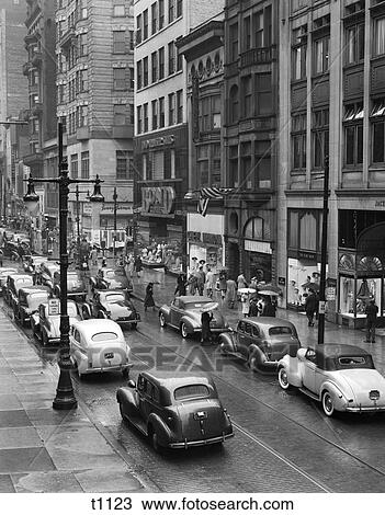 Stock Image - 1940S City Urban Street Scene Rain Wet Stores Parked Cars Pedestrians . Fotosearch