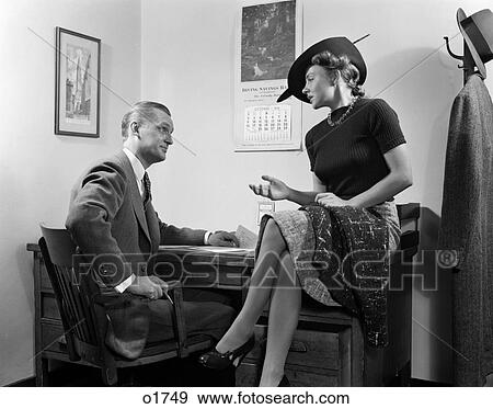Stock Photo - 1940S Couple Man Woman Sitting On Desk Hat Skirt Hand Gesture Talking Discuss Office Secretary Flirt . Fotosearch