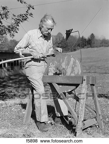Stock Photo - 1940S Elderly Man Seated At Grindstone Outdoors Sharpen Tool Axe Sharp Hone Tool Whetstone Cutting Edge Farmer Tinker. Fotosearch