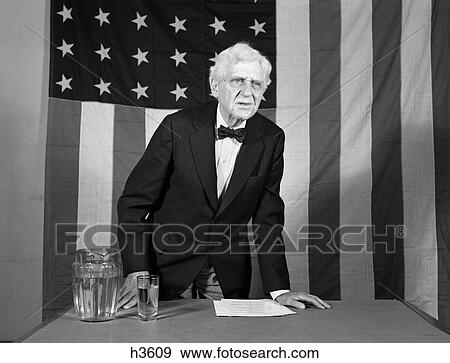 Stock Photo - 1940S Elderly Man Standing At Desk Giving Speech With 48-Star American Flag In Background . Fotosearch