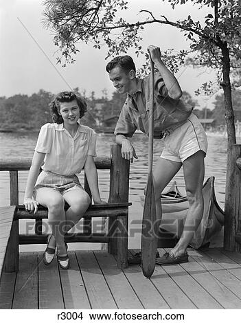 1940S Man And Woman On Dock By Lake Woman Sitting Man Standing Holding Oar Canoe In Background Summer Flirt View Large Photo Image Picture - 1940S Man And Woman On Dock By Lake Woman Sitting Man Standing Holding Oar Canoe In Background Summer Flirt . Fotosearch