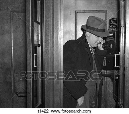 Stock Image - 1940S Man Dressed In A Fedora Suit And Overcoat Standing In A Wood Paneled Phone Booth . Fotosearch