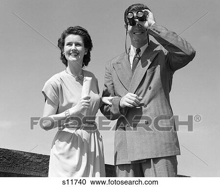 Stock Image - 1940S Smiling Couple Watching A Sporting Event He Is Using A Pair Of Binoculars. Fotosearch