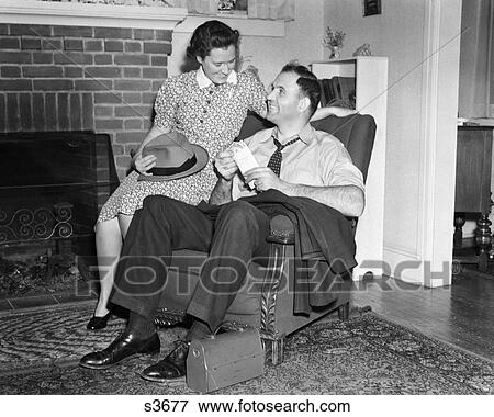 1940S Smiling Man Sitting In Chair Holding Pay Envelope While Wife Sits On Arm Of Chair Holding His Hat Lunch Box Oriental Rug View Large Photo Image Stock Photo - 1940S Smiling Man Sitting In Chair Holding Pay Envelope While Wife Sits On Arm Of Chair Holding His Hat Lunch Box Oriental Rug. Fotosearch