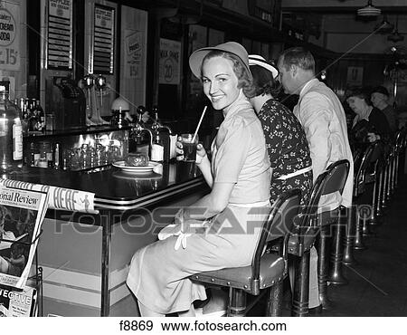 1940S Smiling Women Dressed In A Crisp Outfit & Hat Holding A Glass Of Soda While Sitting At The Counter Of A Soda Fountain View Large Photo Image Stock Photo - 1940S Smiling Women Dressed In A Crisp Outfit & Hat Holding A Glass Of Soda While Sitting At The Counter Of A Soda Fountain . Fotosearch