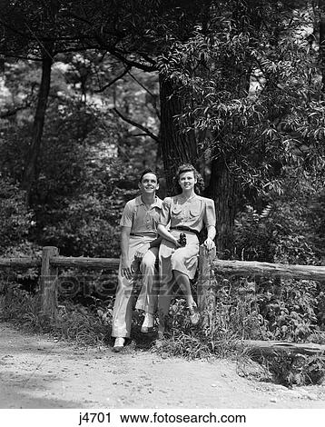 Stock Image - 1940S Teen Couple Sitting On Fence In Woods Looking Up Female Holding Camera . Fotosearch