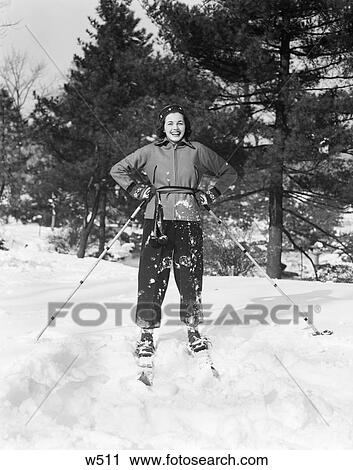 Stock Image - 1940S Woman On Skis Hands On Hips Holding Ski Poles On Ski Slope Slope With Pine Trees In Background Smiling Crisp Fresh Snow. Fotosearch
