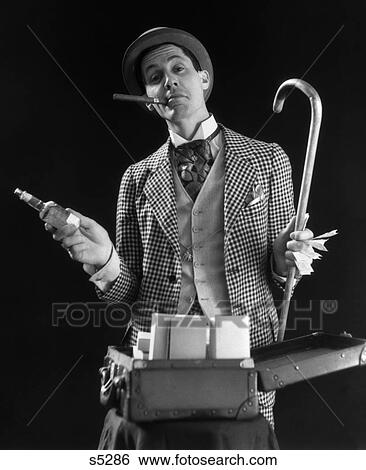 Stock Photograph - Carnival Barker Checkered Suit Cigar In Mouth Holding Cane Money In One Hand Bottle Of Tonic In Other Open Suitcase Stand 1930S . Fotosearch