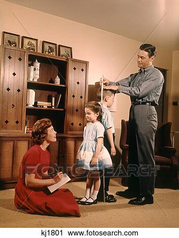 Stock Image - 1950S 1960S Father Mother Son Daughter Measuring Height Sister & Brother Family Indoor Living Room Growing Up . Fotosearch