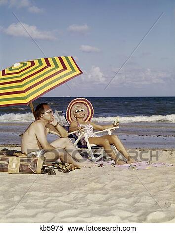 Stock Photography - 1950S 1960S Middle Age Couple Sitting On Beach Under Umbrella Man With Golf Clubs Looks Bored Wife Happy . Fotosearch