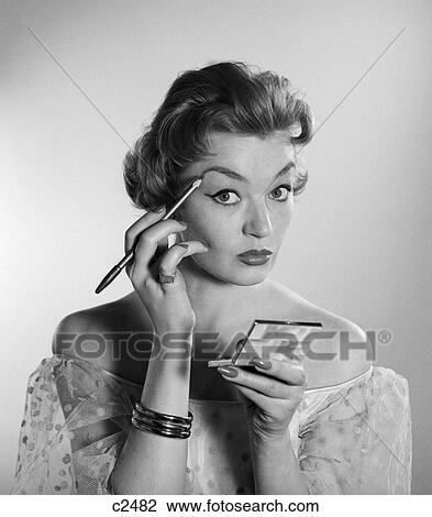 Stock Image - 1950S 1960S Woman Look At Camera Apply Makeup Eye Brow Pencil Holding Compact Arch Beauty Fashion . Fotosearch