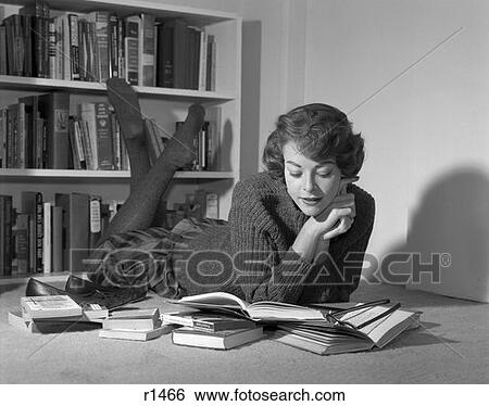 Stock Photograph - 1950S 1960S Young Woman Teen Lying On Floor In Front Of Book Shelves Reading Ankles Crossed . Fotosearch