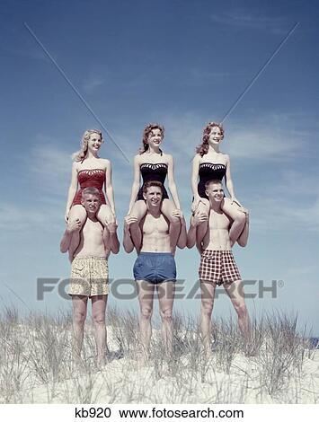 Stock Image - 1950S 3 Teen Couples Stand On Sand Dune Beach Woman Sitting On Mens Shoulders Summer Fun 6 People Piggy Back. Fotosearch