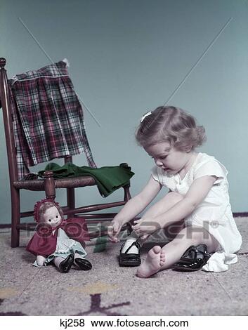 Stock Photo - 1950S Curly Hair Toddler Sit On Floor By Chair And Baby Doll Toy Putting On Her Shoes Patent Leather . Fotosearch