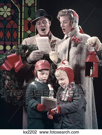 Stock Image - 1950S Family 4 Singing Carols Dad Man Has Wreath Over Arm Mom Woman Holds Red Lantern Boy Girl Music Song Snow Outdoor. Fotosearch