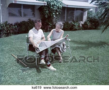 Stock Photo - 1950S Middle Aged Senior Couple Sit In Lawn Chairs In Backyard Rake On Grass Look At Travel Map Retirement Tropical . Fotosearch