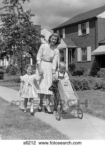 Stock Image - 1950S Mother Walking Down Suburban Sidewalk Holding Daughter'S Hand & Helping Son To Push 2-Wheeled Grocery Cart. Fotosearch