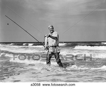 1950S Older Man Standing In Surf In Waders Holding Fish In One Hand Fishing Pole In Other View Large Photo Image Stock Photo - 1950S Older Man Standing In Surf In Waders Holding Fish In One Hand Fishing Pole In Other . Fotosearch