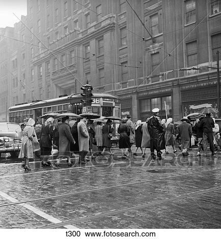 Stock Image - 1950S Pedestrians Intersection City Cross Walk Umbrellas Rain Weather Wet Trolley Car Philadelphia . Fotosearch
