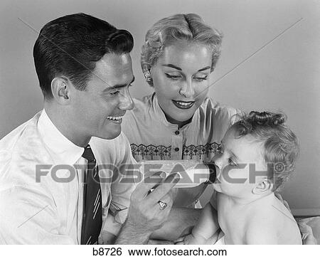Stock Photograph - 1950S Smiling Father Feeding Baby Milk From Bottle With Mother At Side . Fotosearch