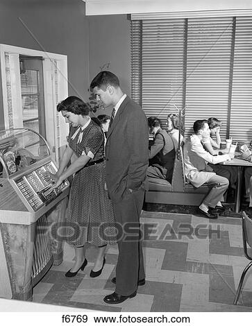 Stock Photo -  1950S Teen Crowd Sitting In Booths With Malts One Couple In Foreground Are Looking At Juke Box The Girl Is Playing It Inside. Fotosearch