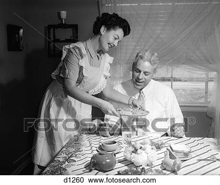 Stock Image - 1950S Wife Wearing Apron Serving Husband Home Cooked Dinner Both Smiling Indoor . Fotosearch