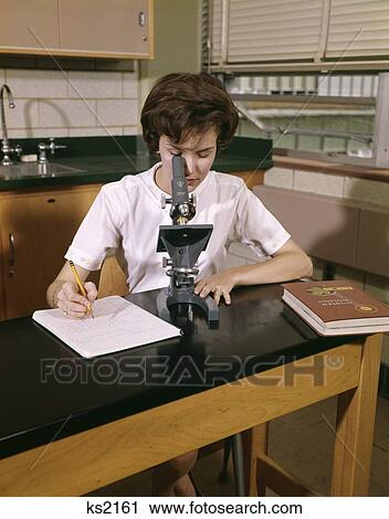 Stock Image - 1960 1960S Female Teen Student Coed At Microscope Taking Notes Biology Textbook High School Retro . Fotosearch