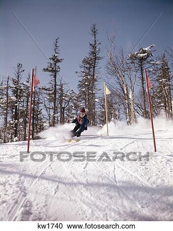 Stock Image - 1960 1960S Man In Blue Outfit Downhill Ski Race Between Flags Gates Speed Sport Skiing Snow Slalom Compete Racing Retro. Fotosearch