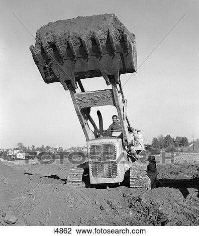 Stock Image - 1960S 1970S Man Operating Caterpillar Heavy Machinery Bull Dozer Lifting Shovel Full Of Dirt Soil Construction Site. Fotosearch