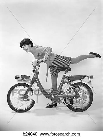 Stock Image - 1960S 1970S Young Woman Smiling Getting On Motor Bike Or Moped With Her Leg Extended Over Back Of Bike . Fotosearch