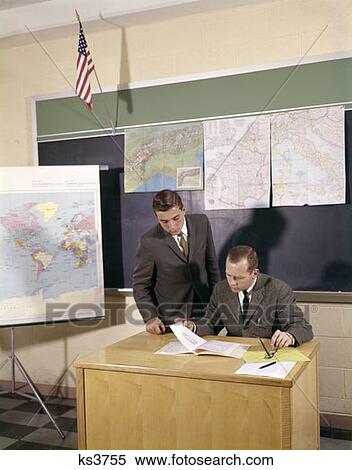 1960s 2 Manner Lehrer Schreibtisch Teenager Schueler Stehen Betrachten Papiere Tragen Anzug Schlips Kreide Ausschuss Karten Fahne Klassenzimmer Stock Fotografie Ks3755 Fotosearch