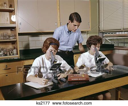 Stock Photo - 1960S 3 Students Biology Classroom Lab Girls Look Into Microscopes Boy Stand Look Over Shoulder Textbook. Fotosearch